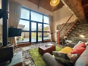 A cozy living room with a stone fireplace, a television, and a large window overlooking a garden. - 1 Bed in Tiverton (oc-96405) (Washfield)