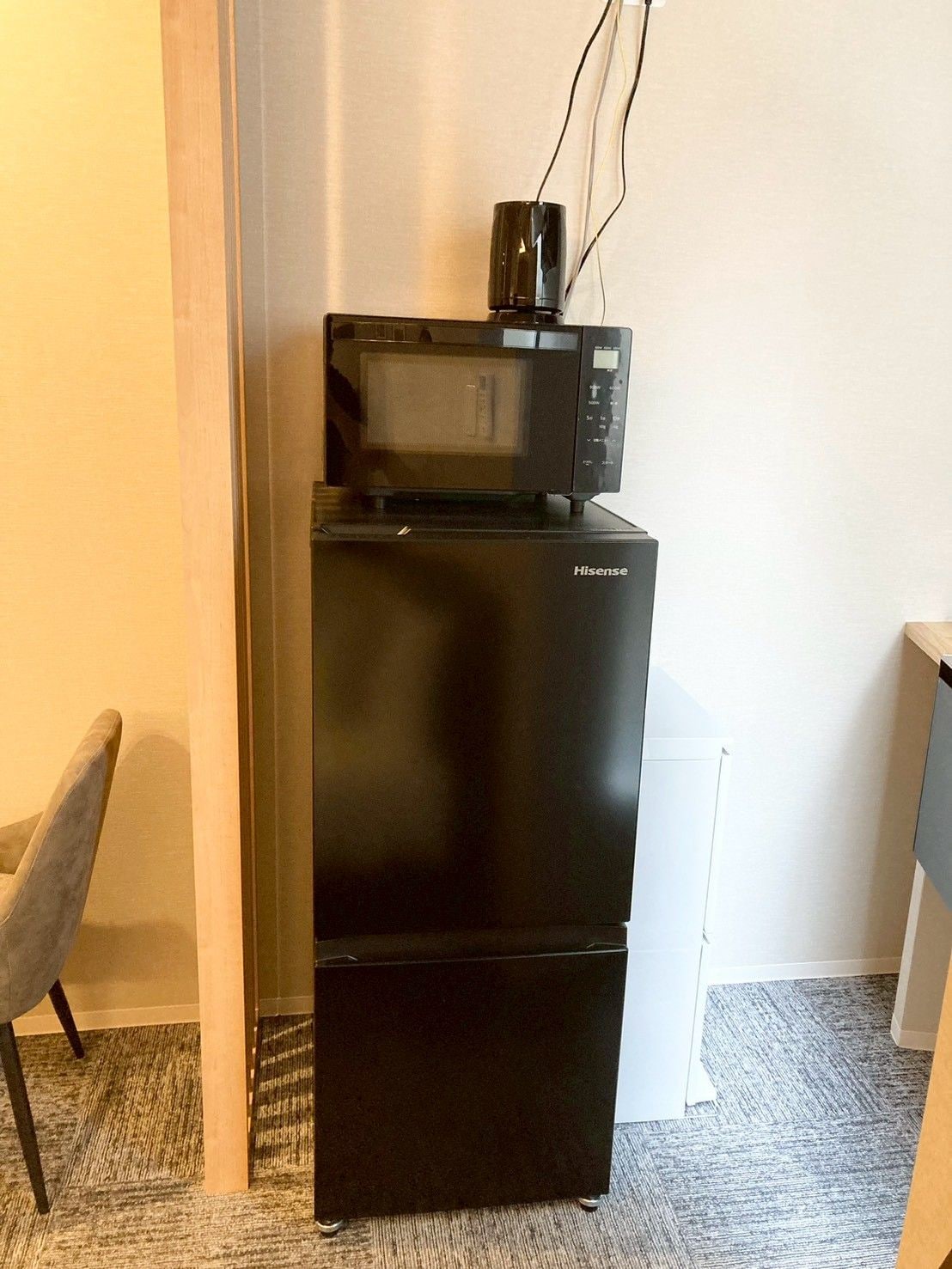 A compact black Hisense refrigerator with a microwave on top, situated in a corner of a room with a carpeted floor and a wooden chair.