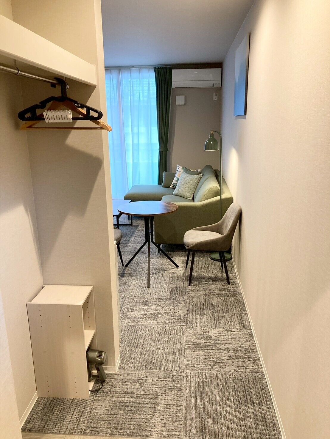 A hotel room with a sofa, a small table, a chair, a hanging coat rack, and a wall-mounted air conditioner.