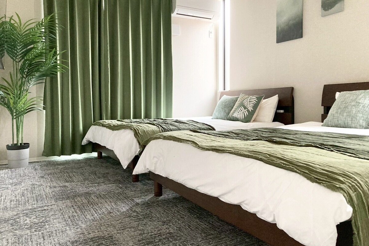 Quarto de hotel com duas camas, uma janela grande com cortinas, um vaso de planta e obras de arte na parede.