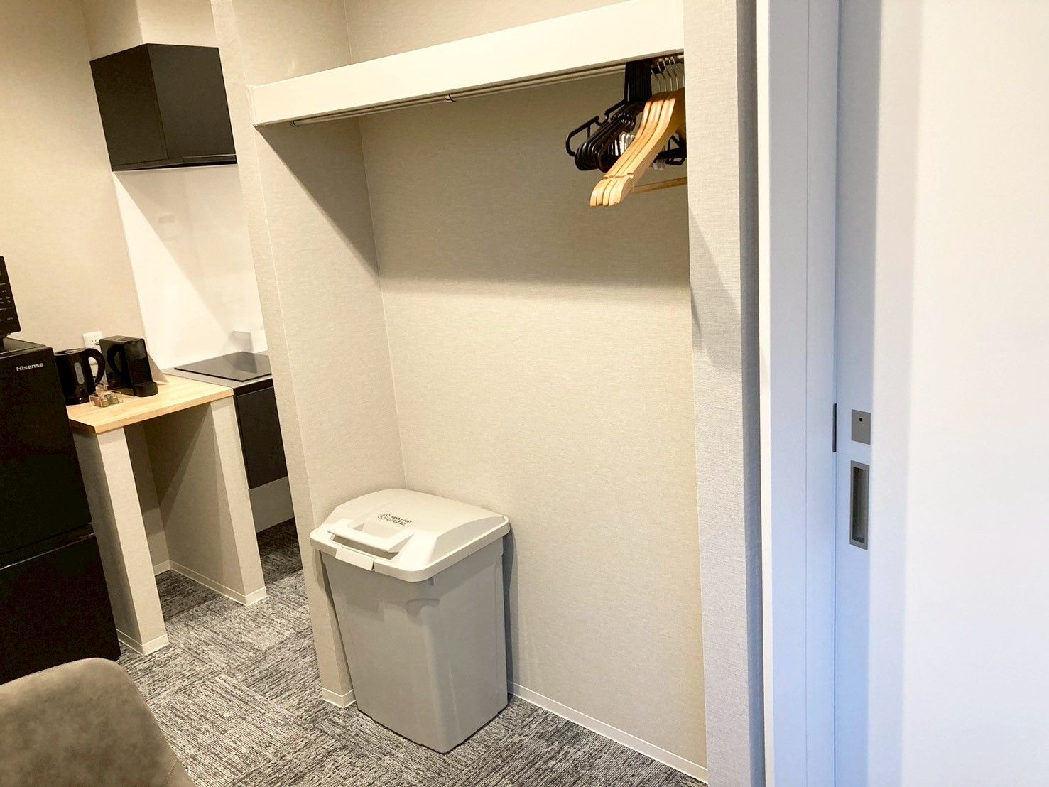 A compact hotel room with a small desk, a trash bin, and a coat rack.
