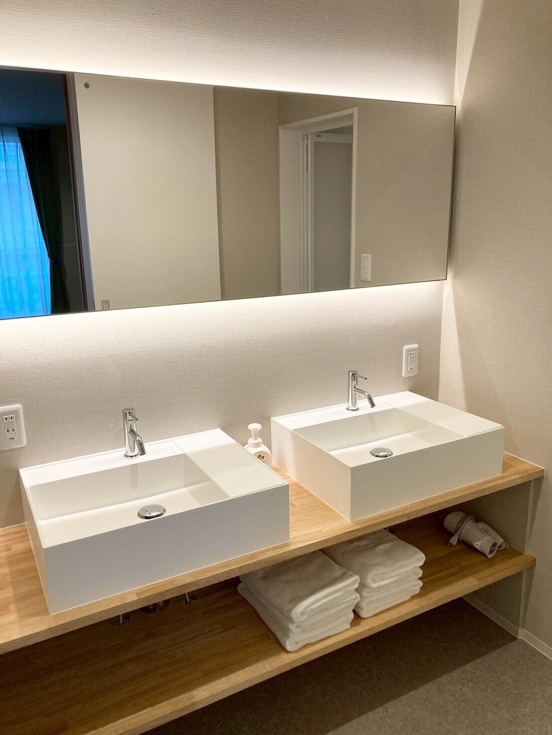 A bathroom with two white sinks, a mirror, and a wooden shelf with towels.