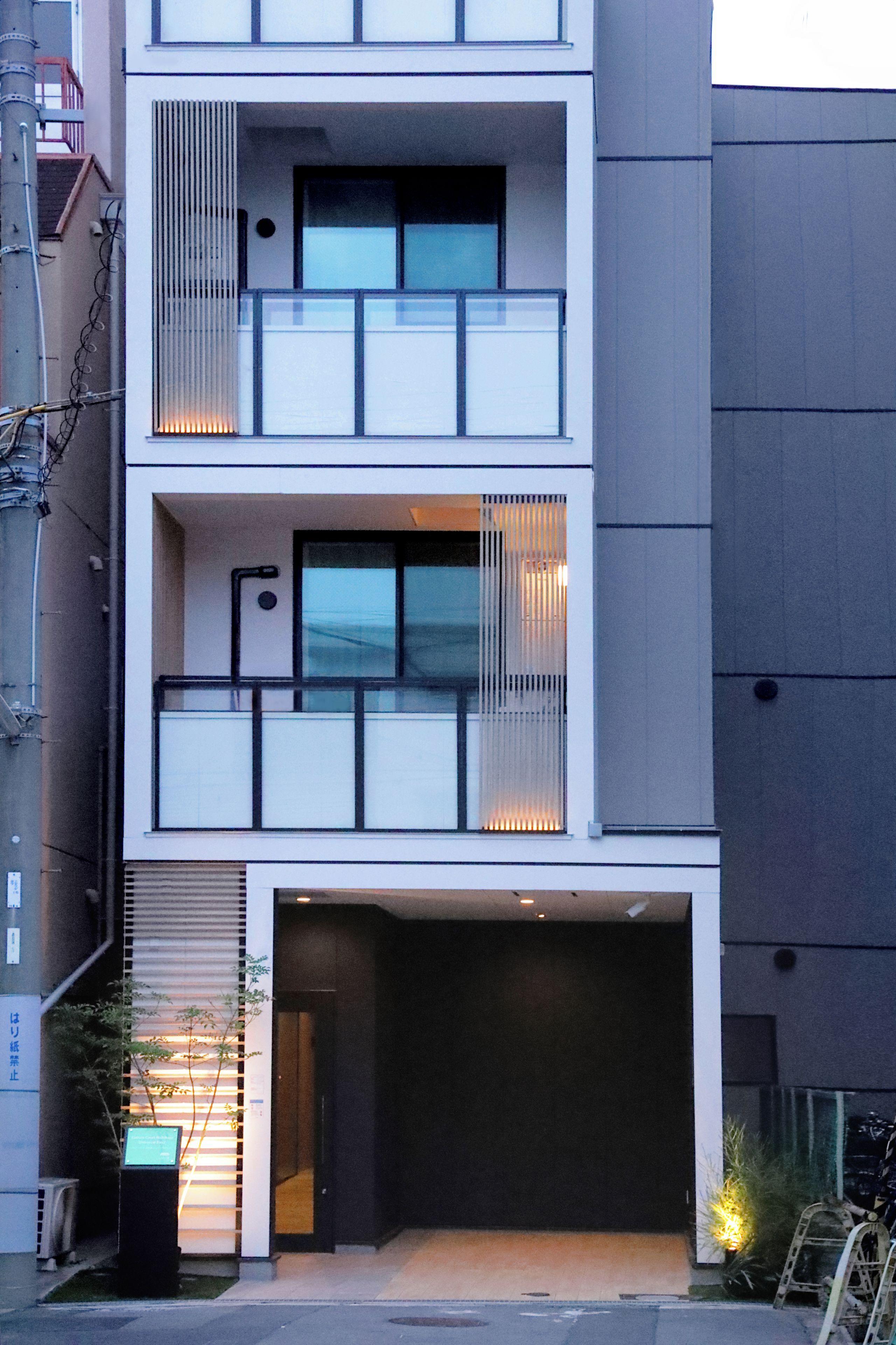 A multi-story building with a garage entrance, a balcony on the upper floor, and a small outdoor area with plants.