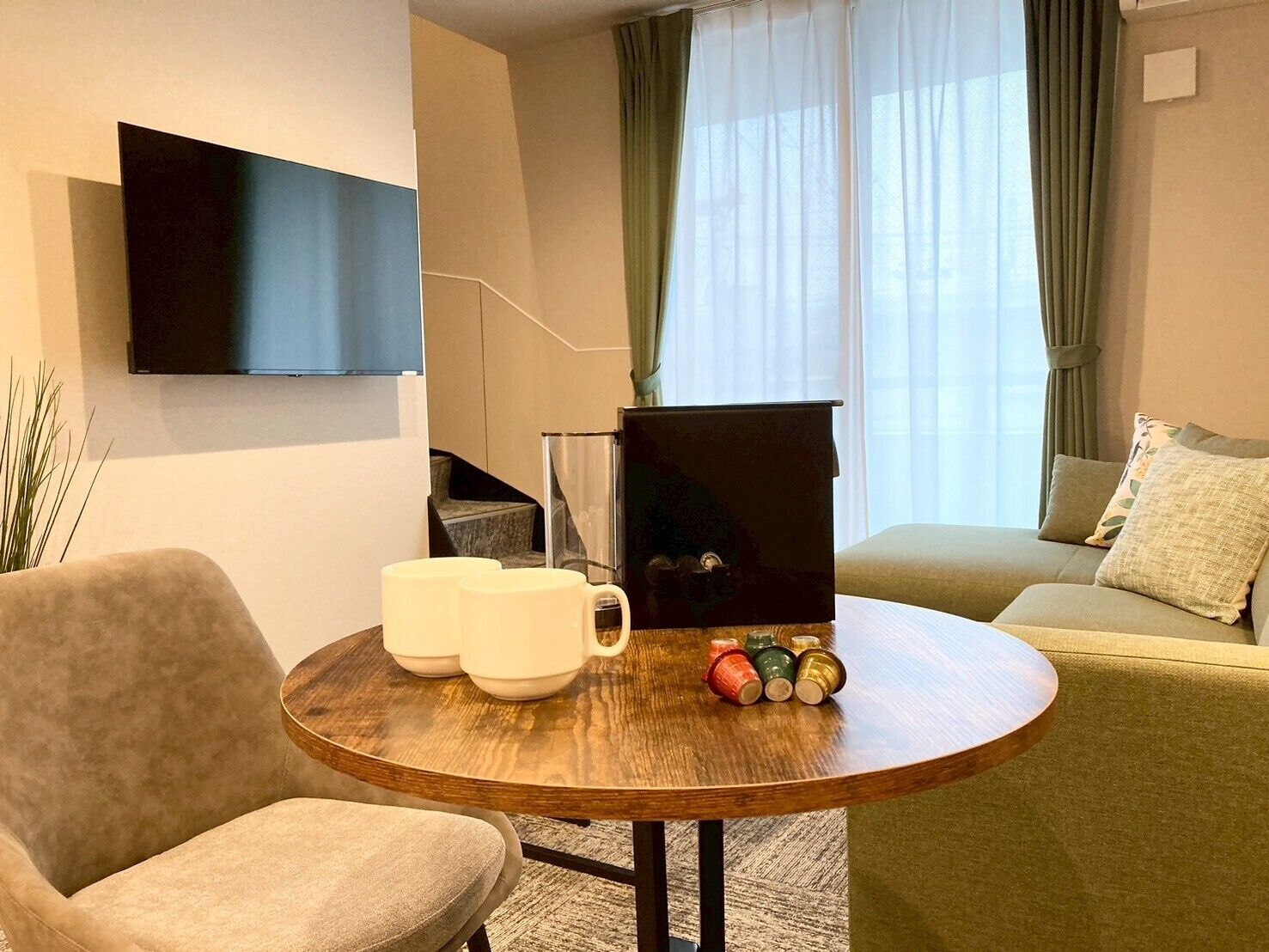 A modern living room with a round wooden table, a sofa, a TV mounted on the wall, and a staircase in the background.