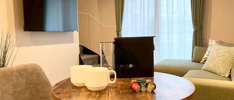 A modern living room with a round wooden table, a sofa, a TV mounted on the wall, and a staircase in the background.