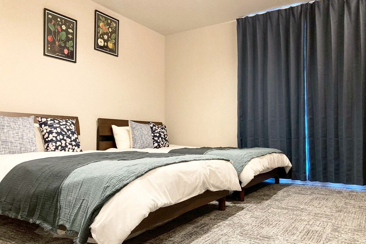 Uma cama de casal com cobertor cinza, duas almofadas, cabeceira e cortina escura.