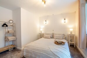 A bedroom with a bed, two bedside tables, a nightstand, a shelf with towels, and a hanging rack.