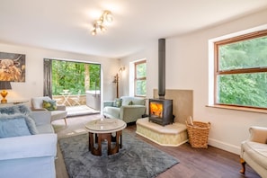 A cozy living room with a fireplace, comfortable seating, and large windows offering a view of greenery.