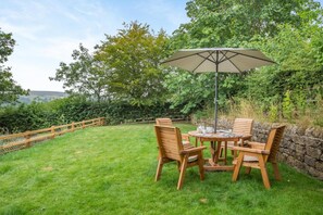 A wooden outdoor table with chairs and a large umbrella, surrounded by a stone wall and lush greenery.