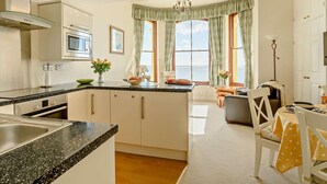 A modern kitchen with a central island, a dining area with a table and chairs, and large windows with curtains. - 2 Bed in Tenby (oc-fb232) (Tenby)