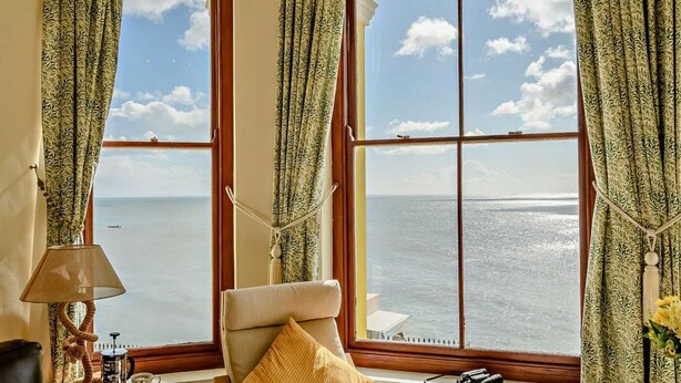 A room with a large window offering a view of the sea, a chair with a cushion, a table with a lamp and a coffee maker, and a decorative rope. - 2 Bed in Tenby (oc-fb232) (Tenby)