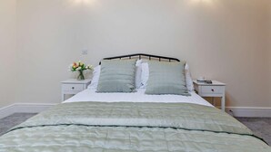 Een net opgemaakt bed met een groene spreidek, twee nachtkastjes met lampen, en een vaas met bloemen op één nachtkastje.