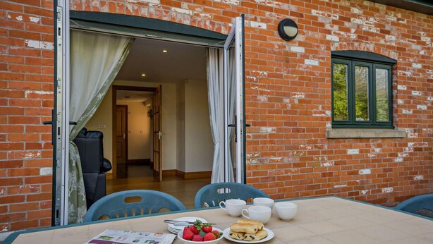 A brick building with a dining area featuring a table with plates of food, cups, and a newspaper. - 5 Bed in Bridport (oc-bigov) (Charmouth)