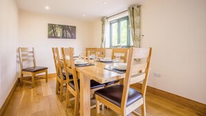 A dining area with a wooden table and chairs, a painting on the wall, and a window with curtains. - 5 Bed in Bridport (oc-bigov) (Charmouth)