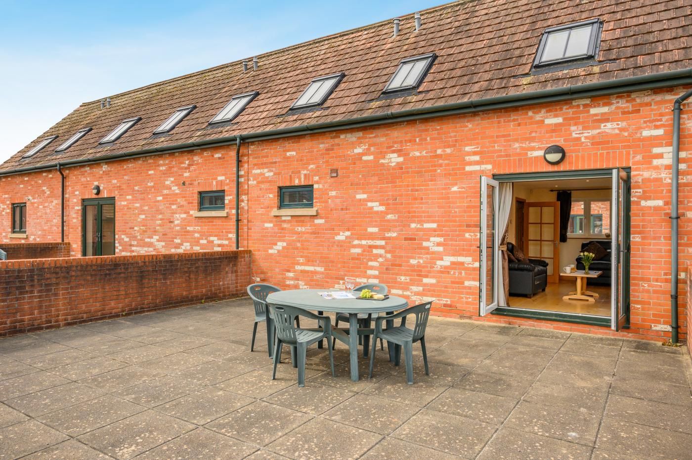 A brick building with a tiled roof, a patio with a table and chairs, and a door leading to an interior space.