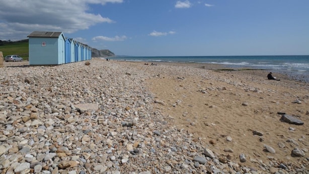 Ein Kiesstrand mit einer Reihe hellblauer Strandhütten und einem klaren Himmel.