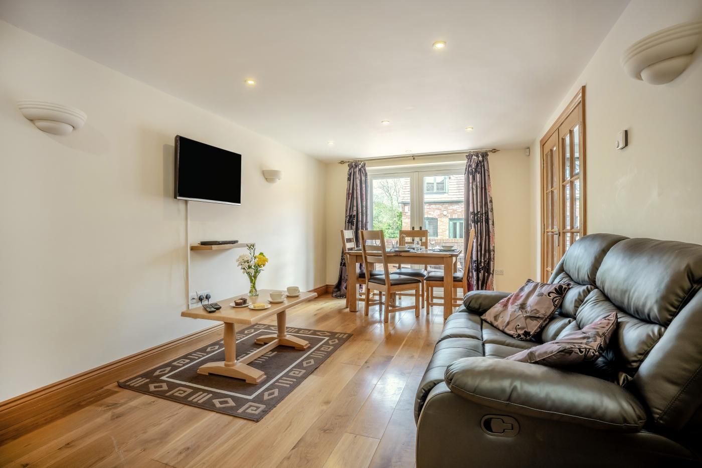 A living room with a leather sofa, a dining table with chairs, a flat-screen TV mounted on the wall, and a window with curtains.