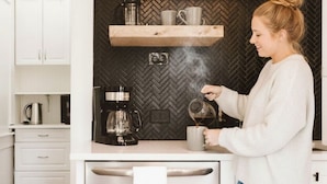 Eine Küche mit weißen Schränken, einer Backspüle mit Fischgrätenmuster, Holzregalen, einer Kaffeemaschine und einer Person, die Kaffee einschenkt.