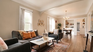 Ein Wohnzimmer mit einer Couch, Sesseln, einem Couchtisch und einem Teppich. Es gibt einen Essbereich mit Tisch und Stühlen.