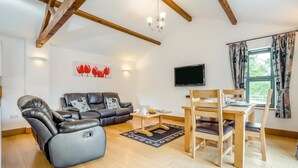 A living room with a leather sofa, a wooden dining table with chairs, a flat-screen TV, and a window with curtains. - 2 Bed in Bridport (oc-restw) (Charmouth)
