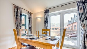 A dining area with a table set for two, a window with curtains, and a view of buildings outside. - 2 Bed in Bridport (oc-restw) (Charmouth)