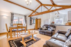 A modern living room with a dining area, wooden beams, a sofa, and a kitchen in the background. - 2 Bed in Bridport (oc-restw) (Charmouth)