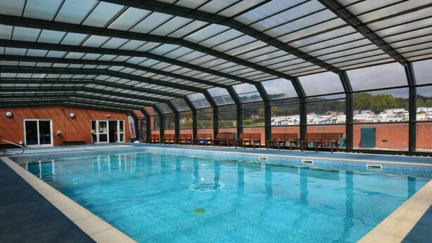 An indoor swimming pool with a glass ceiling, surrounded by brick walls and benches. - 3 Bed in Bridport (oc-tcsaila) (Charmouth)