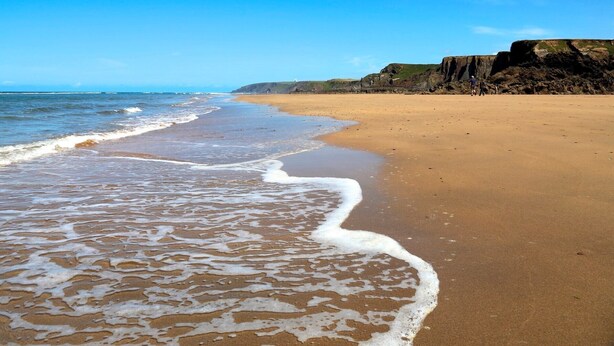 Ein Strand mit Wellen, blauem Himmel und felsigen Klippen.
