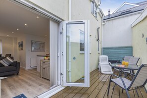 Eine Terrasse mit Tisch und Stühlen, eine Schiebetür zu einem Wohnbereich im Innenbereich und ein kleiner Garten mit einer Bank.