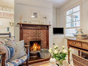 Ein gemütliches Wohnzimmer mit Kamin aus Ziegelsteinen, einer Couch mit floralen Kissen, einem Couchtisch und einer Vase mit weißen Blumen.