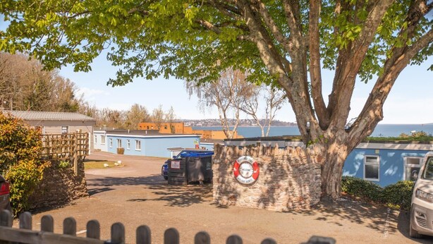 Une propriété située en bord de mer, dotée d’un espace de stationnement clôturé, d’un panneau indiquant la présence d’une bouée de sauvetage et offrant une vue sur la mer.