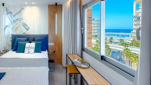 Une chambre d’hôtel moderne avec un grand lit, une table de chevet en bois, un banc et une vue sur le port de plaisance et les bâtiments.