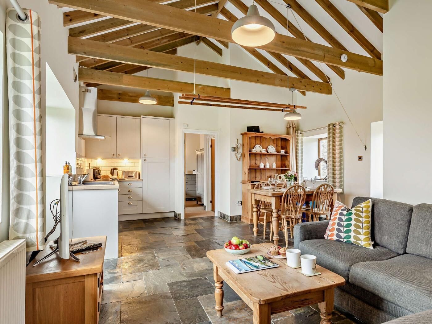 A modern kitchen with white cabinets, a dining area with wooden chairs, and a grey sofa with patterned cushions.
