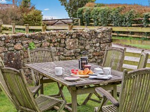 A rustic outdoor dining setup with a stone wall, wooden chairs, and a table set with breakfast items. - 2 Bed in Bude (oc-04727) (Poundstock)