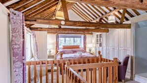 Ein Schlafzimmer mit Holzbalkendecke, einem Bett mit Blumenmuster-Überwurf, einem Stuhl und einem Fenster mit Vorhängen.