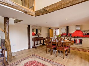 Un grand espace à manger avec une table et des chaises en bois, un tapis aux motifs rouges et beiges, et une cuisine dont le mur du fond est recouvert de carreaux rouges.