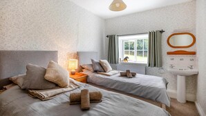 A bedroom with two beds, a bedside table with a lamp, a window with curtains, a radiator, and a sink with a mirror. - 3 Bed in Barnstaple (oc-wayco) (Shirwell)