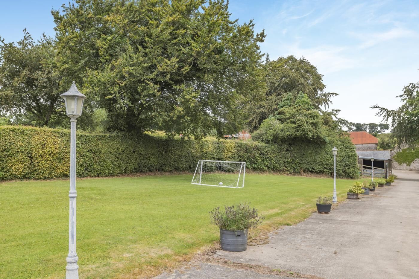 A well-maintained lawn with a soccer goal, surrounded by trees and a hedge, with a pathway leading to a building.