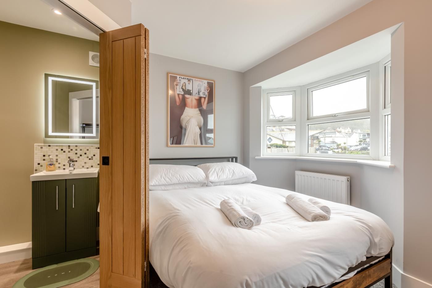 Un dormitorio con cama, ventana, baño con espejo y una revista en la pared.