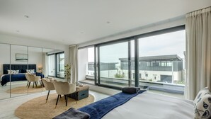 Una habitación de hotel moderna con una cama grande, un área de descanso con sillas, un ventanal con vista a la ciudad y un espejo.