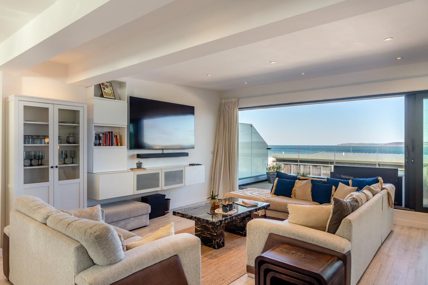 A modern living room with a large TV, comfortable seating, a bookshelf, and a balcony with a view of the sea.