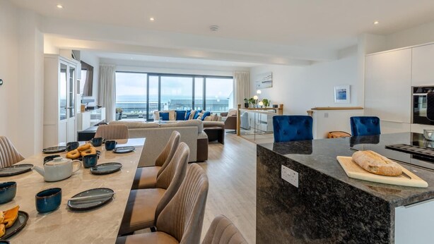 Una cocina moderna con encimera de mármol, un comedor con una mesa larga y sillas, y una sala de estar con sofá y un amplio ventanal con vistas a la ciudad.