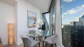 Een moderne kamer met een glazen tafel, twee stoelen, een schilderij en uitzicht op een stadsbeeld door grote ramen.
