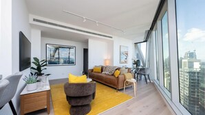 Een moderne woonkamer met een bank, fauteuil en eethoek, met grote ramen en kunstwerken aan de wanden.