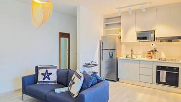 A modern apartment with a grey sofa, a kitchen with white cabinets, a stainless steel refrigerator, and a hanging light fixture.