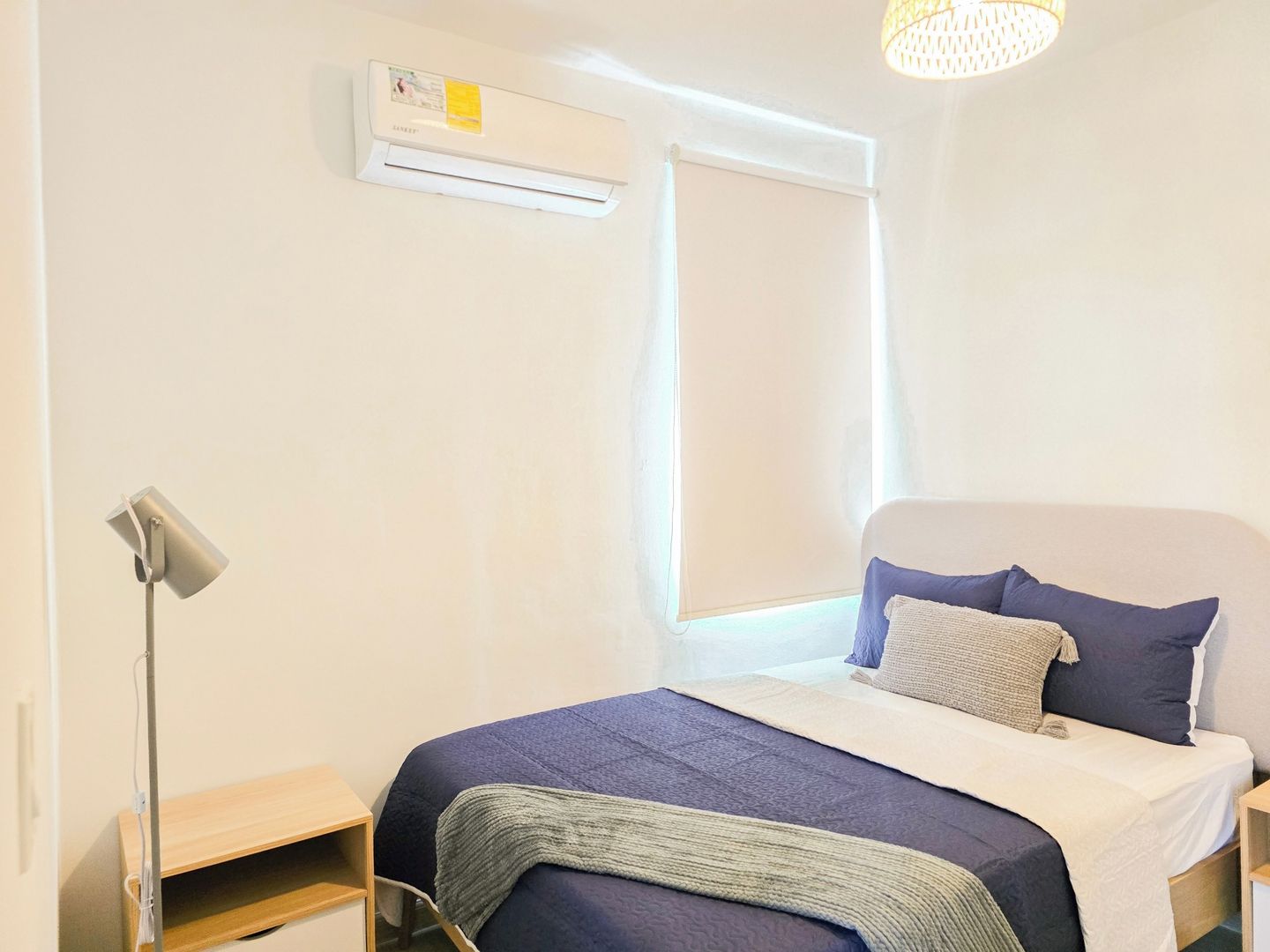 A bedroom with a bed, a bedside table, a lamp, an air conditioning unit, and a window with a blind.