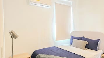 A bedroom with a bed, a bedside table, a lamp, an air conditioning unit, and a window with a blind.