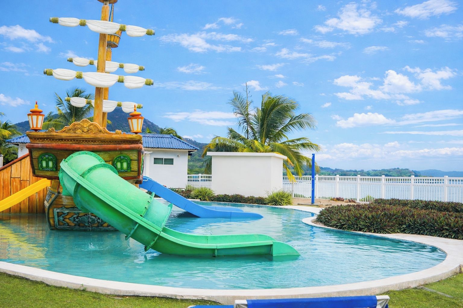 A pool with a green slide, a ship-themed play structure, and a white building in the background.