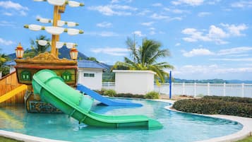 A pool with a green slide, a ship-themed play structure, and a white building in the background.