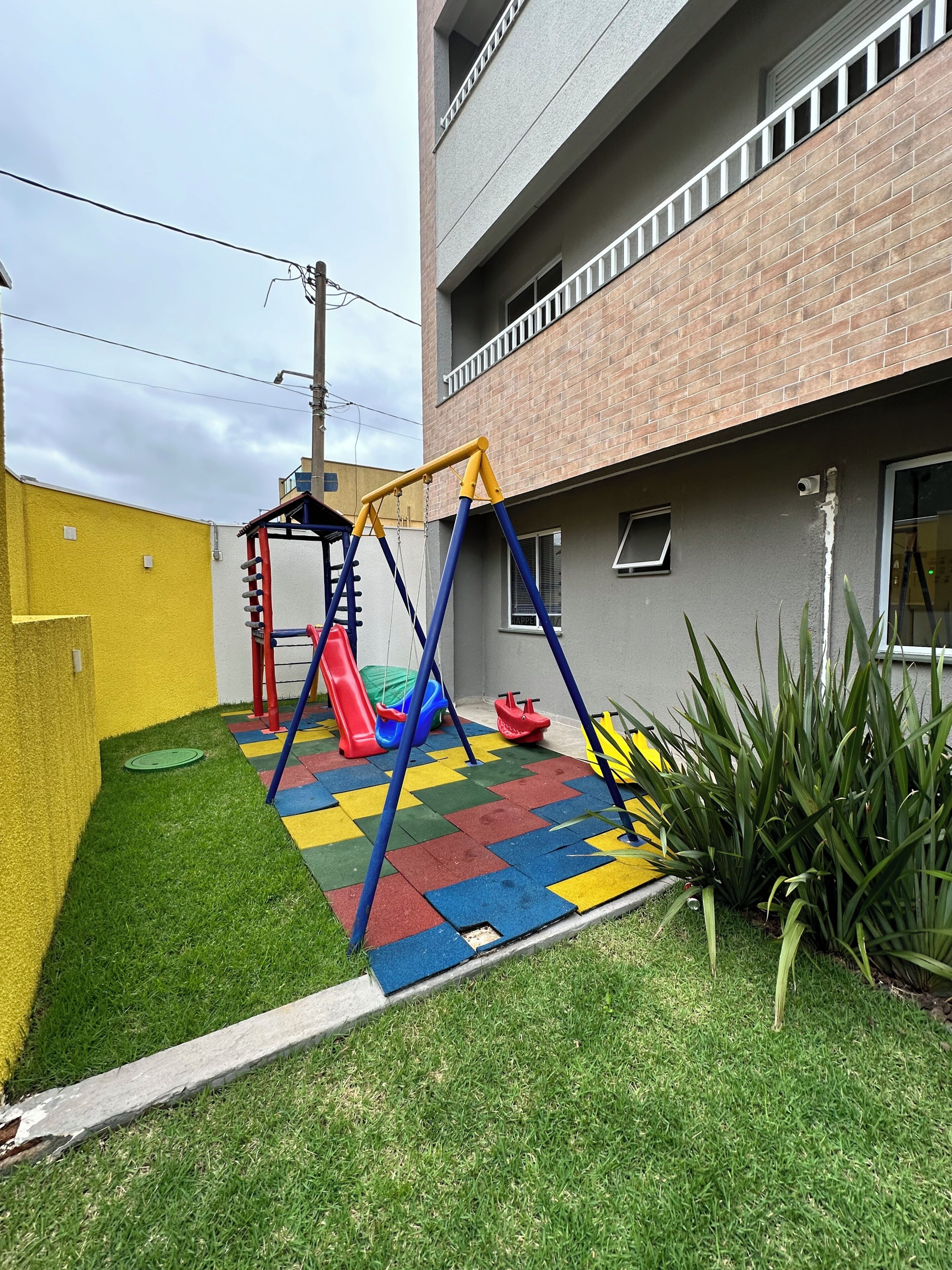 Une aire de jeux avec des balançoires et des toboggans, un mur jaune et un bâtiment à la façade en briques.